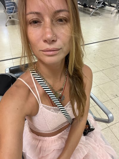 A woman with long, blonde hair and a gold nose ring is sitting in an airport waiting area. She is wearing a pink lace bralette and a pink, layered skirt. A black and white striped strap is visible across her chest, and she has several necklaces on. She is holding a small black bag with a multi-colored strap. The background shows rows of grey airport chairs and tiled flooring. The scene appears to be the Bandaranaike International Airport in Katunayake, Sri Lanka.
