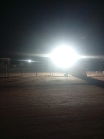 The image depicts an outdoor scene at night, illuminated by a bright, overhead light source. The light is so intense that it washes out much of the detail in the central area of the image. In the foreground, there is a flat, dusty or sandy ground.
Behind the illuminated area, a fence or barrier is visible, suggesting some sort of enclosure or designated space. Beyond the fence, there appear to be structures, possibly buildings or stalls, dimly lit or obscured by the glare. A cylindrical object, likely a trash can or bin, stands near the center of the image. The overall setting appears to be an open area, possibly a marketplace, a roadside stop, or a service station, given the structures and the presence of what might be a sign on the left side, though any text is illegible.
The time of day is clearly night, and the weather appears to be clear, with no visible rain or fog. The bright light creates long shadows, indicating a single, powerful light source from above. There are no people visible in the frame. The mood is somewhat stark due to the high contrast lighting and lack of discernible activity. The location context of Tundun Wada South, Nigeria, suggests a potential urban or semi-urban environment within that region.