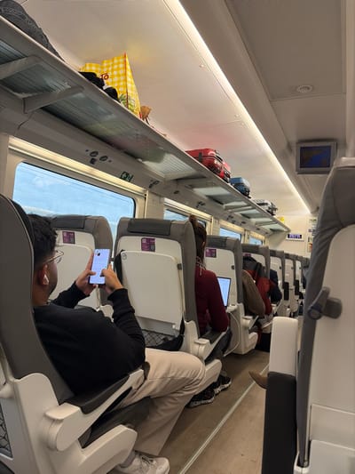 The image displays the interior of a modern passenger train, likely in motion, as observed from a seat in Alcalà de Xivert, Spain. The scene shows multiple passengers seated along a single aisle. In the immediate foreground, a male individual is seated facing forward, wearing a black long-sleeved shirt, light-colored trousers, and white sneakers. He has dark hair, wears glasses, and has white earbuds in his ears. He is actively holding and interacting with a white smartphone. Directly in front of him, a female passenger with dark hair tied up is seated, wearing a dark red long-sleeved top and using a silver laptop positioned on her tray table.

The train car features rows of grey and white passenger seats with built-in tray tables and mesh pockets on the seatbacks. Overhead luggage racks extend along both sides of the car, containing various items including a large yellow-and-white checkered bag, a black backpack, a red hard-shell suitcase, a blue hard-shell suitcase, and several other bags. Large rectangular windows line the side of the train, revealing a bright, overcast sky outside. The ceiling is white with integrated linear lighting and air vents. Towards the rear of the visible section on the right, a small screen displays a map, likely a route or location indicator. Emergency exit signs are visible above the windows. Several other passengers are seated further down the aisle, also facing forward. The floor is a light grey with a darker central strip.