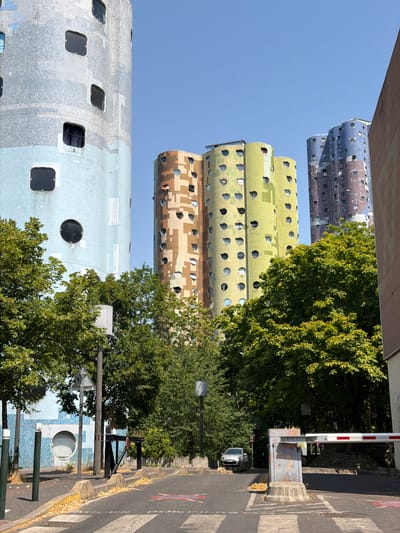 This media file captures a street-level view of residential buildings in Nanterre, France. The buildings, characterized by their cylindrical shape and varied window patterns, are a prominent subject. One building displays a mosaic of blue and silver, with square and circular windows. Another features a camouflage-like pattern of brown and beige with numerous circular windows. A third, further back, is predominantly yellow with similar circular windows. Trees with lush green foliage are interspersed between the buildings and line the street, suggesting a temperate climate and possibly springtime or summer.
The scene appears to be during the daytime, indicated by the clear blue sky and bright sunlight casting shadows. The presence of a pedestrian crossing, a barrier arm, and a parked car on the right side of the image suggests an urban or suburban residential area. A street lamp and a traffic sign are also visible. There is no discernible activity or event taking place; the image seems to be a static capture of the architecture and surroundings. No text is visible in the image. The overall impression is one of a unique architectural style within a green, everyday setting.