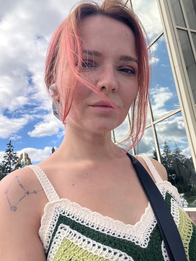 A close-up, waist-up, selfie taken outdoors on a sunny day. The subject, a young woman with pink-dyed hair and freckles, is looking towards the camera. She is wearing a crocheted top with white and green patterns and a black strap. A small tattoo of a constellation is visible on her left shoulder. The background includes a blue sky with white clouds, trees, and a modern building with reflective glass windows. The environment suggests an urban setting, potentially near a park or a public building in Mezhdurechenskiy, Russia.