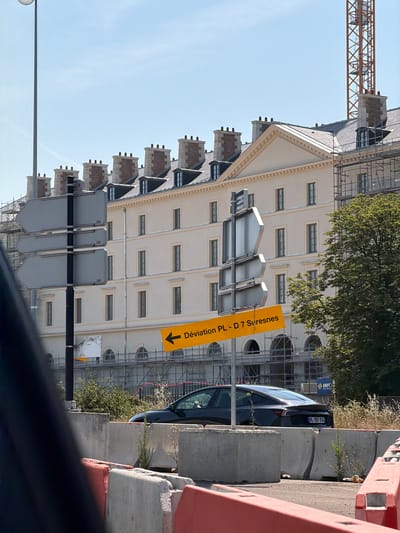 A dark colored car is parked behind a concrete barrier in Boulogne-Billancourt, France. The car has a license plate that reads "HL-397-EX". In the background, a light colored building with many windows and chimneys stands against a clear blue sky. A large orange construction crane is visible behind the building. A yellow directional sign with black text reads "Déviation PL - D 7 Suresnes" and points to the left. There are multiple road signs on a black pole in front of the building. The scene appears to be a city street with ongoing construction.