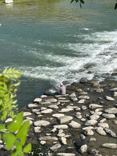 The image shows a man fishing in a river in Buk-myeon, South Korea. The river flows from left to right, with a rocky bank on the right side. The man is wearing a grey shirt and a pink hat, and is standing on the rocky bank, holding a fishing rod. The water is clear and flowing rapidly. The environment includes rocks and vegetation.