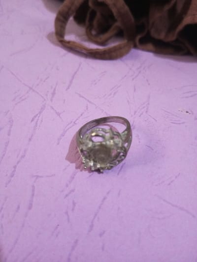 A silver-toned metallic ring is positioned on a textured, light purple surface in Chandragiri, Nepal. The ring features an open-work, ornate setting designed for a central gemstone, which is currently absent; small prongs are visible around the empty bezel. A distinct shadow extends from the ring towards the lower-left. In the upper right background, a crumpled section of dark brown fabric or material is partially visible.