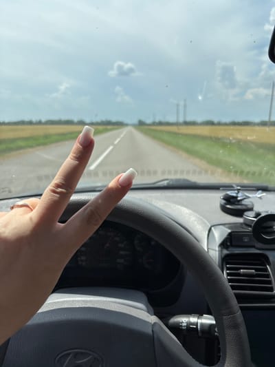 The image depicts a first-person perspective from inside a vehicle, showing the driver's hand making a peace sign over the steering wheel. The road ahead is visible, stretching into the distance through a flat, rural landscape characterized by fields of golden crops and green vegetation. The sky is partly cloudy, with a bright, hazy sun. Inside the car, a portion of the dashboard is visible, including a car's steering wheel, and a small toy helicopter is perched on the dashboard. The location is not specified within the image, and no information about Maryinskiy, Russia, can be deduced.
