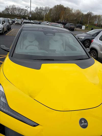 The image depicts a bright yellow Lotus Emira parked in a car park in Braintree, United Kingdom. The vehicle is angled in the frame, showing the front right portion of the car. The Lotus logo is visible on the right front panel. Other vehicles are parked in the background. The sky is overcast. Trees line the background behind the parking area.