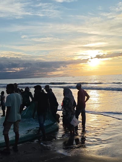 In the city of Uleekareung, Indonesia, a group of people are gathered on a sandy beach at sunset. The sky is filled with dramatic clouds in shades of blue and orange, illuminated by the setting sun. The ocean is calm with gentle waves lapping at the shore. The people appear to be fishermen, as some are seen pulling a large fishing net from the water. The mood is one of activity and communal effort. The overall scene is serene yet dynamic, capturing a moment of daily life in this coastal Indonesian community.