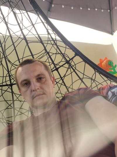 This is a close-up, low-angle selfie of a man's face and upper body, taken outdoors. He is looking directly at the camera with a neutral expression. His short brown hair is slightly messy. He is wearing a maroon t-shirt.
The man is positioned in front of a black, woven structure, possibly a chair or a hammock, which forms a complex, intersecting pattern in the foreground. Behind him, a large, dark object with visible silver studs, likely an umbrella or canopy, is partially visible on the left. The background is bright, suggesting daylight, with a pale, possibly wall-like surface. A colorful toy, shaped like an orange flower with green accents, is also visible in the upper part of the image, hanging from the woven structure. The overall lighting is bright and appears to be natural sunlight, indicating it is daytime. The scene suggests a casual outdoor setting, possibly a patio or backyard in Švenčionys, Lithuania. There is no visible text in the image. The motion blur in some parts of the image indicates movement during the photograph.