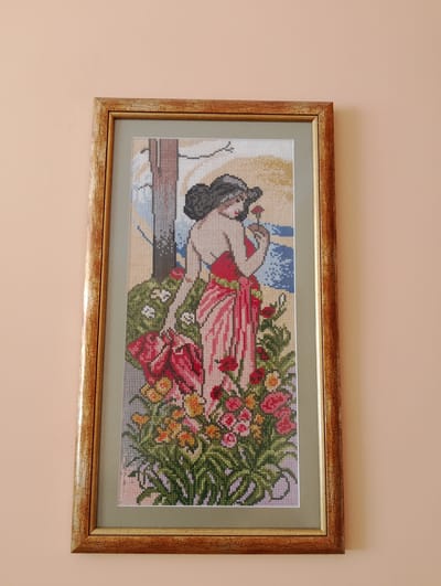 A framed cross-stitch artwork hangs on a peach-colored wall. The artwork depicts a woman in a red dress standing amidst a vibrant garden of flowers. She has dark hair styled in an updo and is holding a single red flower to her nose. In her left arm, she carries a red cloth bag or scarf. Behind her, a suggestion of a tree trunk and a hazy, light-colored background with hints of blue, possibly representing water or sky, can be seen. The flowers in the foreground are a mix of red, pink, yellow, and white, with abundant green foliage. The cross-stitch technique is evident in the pixelated appearance of the image. The frame is made of wood with a textured, golden-brown finish, showing some cracking and wear. The overall impression is one of a serene moment in nature. The image does not contain any visible text. The location context is Kolno, Poland.