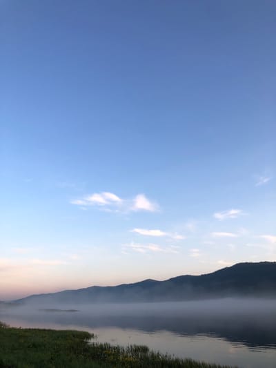 The image depicts a serene natural landscape at dawn or dusk. A calm body of water, likely a river or lake, occupies the lower half of the frame. A gentle mist hovers over the water's surface, obscuring the base of distant, tree-covered mountains that form a dark silhouette against the sky. The sky is a gradient of soft blues and pinks, with a few wispy clouds scattered across it. In the foreground, the edge of the water is lined with lush green grass, interspersed with small yellow wildflowers. The scene is devoid of any human presence or man-made structures, suggesting a remote, undeveloped natural setting within or near the city of Sliznevo, Russia. The overall atmosphere is peaceful and tranquil.
