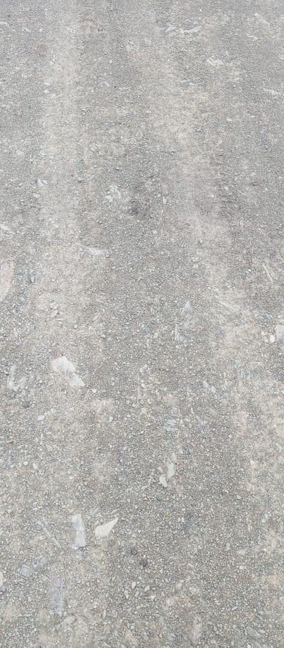 The image displays a close-up, overhead view of a gravel surface. The ground is composed of small, irregularly shaped grey and white pebbles and stones, interspersed with finer gravel and dirt. Some larger, flatter stones are visible, some appearing lighter in color. The surface is uneven and appears to be outdoors. The context of Khusainovo, Russia, is a designation of location and does not influence the visual description of the ground. There are no people, animals, or specific objects of note other than the natural composition of the ground itself. The environment is characterized by the rough, textured surface of the gravel.