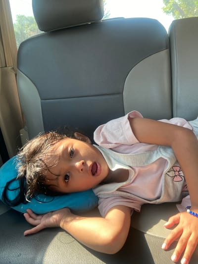 A young child with dark hair lies on a blue pillow in the back seat of a vehicle. The child is wearing a pink t-shirt and overalls with a bow design. Sunlight streams in from the right side of the image, casting shadows on the child and the gray and tan leather seats. The background shows trees and sky through the vehicle window. The location is Ngrame, Indonesia.