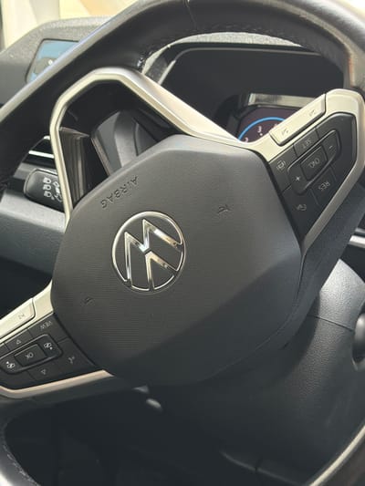 The image shows a close-up view of a car's steering wheel. The steering wheel is black with silver accents and features various control buttons. Prominently displayed in the center of the steering wheel is the Volkswagen logo. To the left of the logo, the word "AIRBAG" is visible.
On the left side of the steering wheel, there are buttons labeled "SET", "CNCL", and "RES". There are also buttons with plus and minus symbols, as well as a button with an "i" symbol. On the right side of the steering wheel, there are buttons labeled "VIEW" and "OK", along with directional arrows.
The background of the image is dark, suggesting the interior of a car. A portion of the car's dashboard is visible on the left side, showing a digital display with some blue and white markings. The image appears to be taken in daylight, as indicated by the lighting. There are no people visible in the image. The setting is likely a parked car.
The visible text includes "AIRBAG", "SET", "CNCL", "RES", "VIEW", and "OK". There are also some symbols on the buttons, including plus, minus, and directional arrows. The Volkswagen logo is also a recognizable symbol. The overall impression is of a modern car's interior, focusing on the driver's controls.