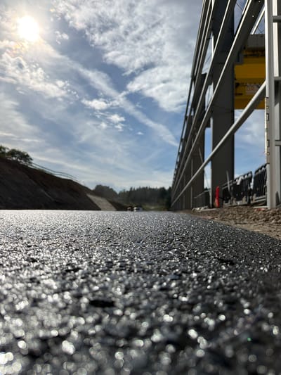 The image shows a low-angle perspective of a newly paved asphalt surface, likely a road or construction site, in Janowice, Poland. The asphalt is glistening with sunlight, creating numerous bokeh effects in the foreground. In the middle ground, a construction site is visible with a large yellow crane and grey metal framing. A sloped embankment covered in dark soil is on the left, with trees and foliage in the distance. The sky is blue with scattered white clouds and the sun is bright and partially obscured by clouds. The scene appears to be a construction project in progress.