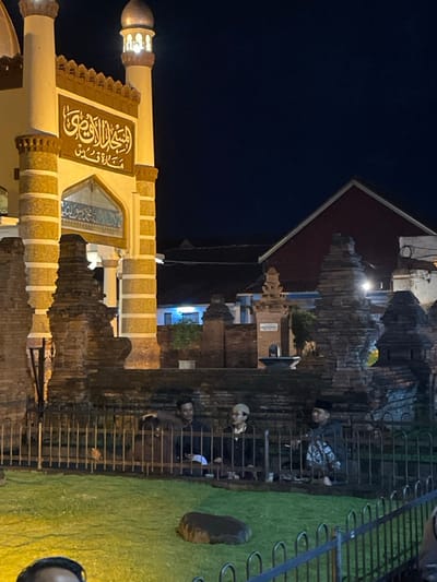 The image depicts a nighttime scene in Kauman, Indonesia, featuring a brightly lit mosque entrance and minaret on the left, set against a dark sky. The mosque structure presents light-colored pillars with horizontal banding and dark reddish-brown accents. An arched entryway is visible, topped with light blue tilework and a rectangular panel featuring prominent dark Arabic calligraphy on a light background. An illuminated, dome-topped minaret rises on the far left.

In the mid-ground, a neatly maintained green grass lawn occupies the lower portion of the frame, separated from the viewer by a dark metal fence with vertical bars. A dark, irregularly shaped object rests on the grass. Behind this fence, on a slightly elevated area, approximately four individuals are seated or crouching. They wear dark casual clothing; one person wears a white skullcap, and another a black peci. Their specific actions are not clearly discernible, but they appear to be engaging in quiet activity or conversation. The upper head of a fifth person is partially visible in the bottom-left corner, outside the fence.

Multiple weathered, dark reddish-brown brick structures, exhibiting a stacked or tiered design, are positioned between the grass area and the mosque, extending towards the right. These structures appear to be part of a surrounding wall or decorative elements. In the background, under the dark sky, several indistinct buildings with dark red-sloped roofs are visible, along with a smaller light-colored structure featuring a tower-like element near the center-right. The scene is illuminated by artificial light sources, casting a warm glow on the foreground and mosque.