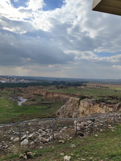 The image presents a panoramic view of a landscape identified as being in Rosh Haayin, Israel. The scene is observed from an elevated position, with a portion of a light-colored overhead structure visible in the upper right corner.

The sky occupies the upper half of the image, dominated by large, varied grey and white clouds, with interspersed patches of blue sky visible. Faint light rays penetrate the cloud cover in the distant background.

In the foreground, a gentle slope covered with sparse green grass and numerous scattered white and light-colored rocks descends from the viewer's perspective. A grey metal barrier, consisting of vertical posts and horizontal rails, runs horizontally across the lower third of the frame, delineating the edge of the elevated viewpoint.

Immediately beyond the barrier, the midground reveals a large excavated area, consistent with a quarry. This area features prominent, multi-layered rock cliffs and steep earthen embankments of a light brown and beige hue. A sinuous body of water, resembling a pond or a segment of a stream, is situated within the lower section of this quarry, bordered by dense green marshland vegetation. Additional green grassy patches are visible on and around the rock formations.

The background extends into a flat, expansive plain. This landscape consists of cultivated agricultural fields displaying varying shades of green, interspersed with clusters of darker green trees. In the far distance, a hazy urban area with discernible buildings is visible, consistent with the city of Rosh Haayin or its immediate vicinity. No individuals or specific activities are depicted in the scene.