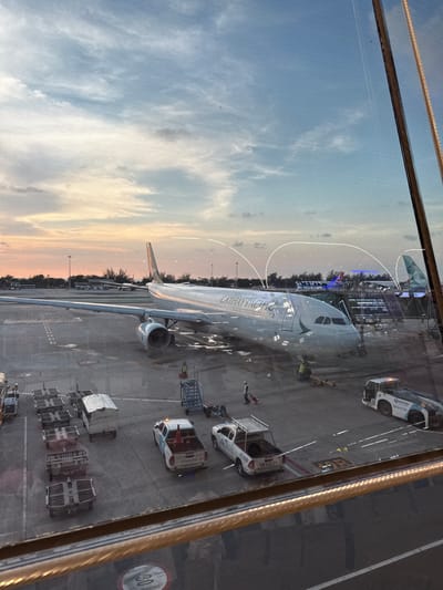 View through a window of a Cathay Pacific airplane at an airport. Ground crew vehicles surround the plane. The terminal is visible in the background with a blue sign written in Thai script. The sky is visible with pink and orange hues indicating sunrise or sunset. The speed limit sign "30" is visible near the right side of the frame. The reflection in the window adds a layer of visual complexity. The location appears to be Phuket International Airport in ตำบล ไม้ขาว, Thailand.