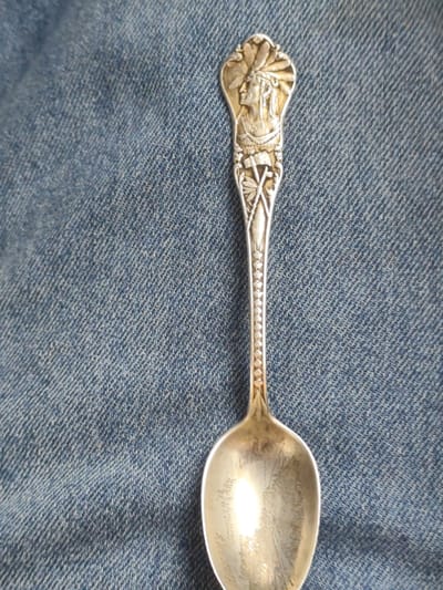 This is a close-up, top-down shot of a silver spoon lying on a blue denim fabric. The spoon features intricate detailing on its handle, depicting a profile of a Native American man wearing a feathered headdress. Below the headdress are crossed tomahawks, with decorative leaf-like elements surrounding them. The handle has a textured, beaded pattern along its edges, tapering down to the bowl of the spoon. The bowl itself is somewhat tarnished and reflects light. Visible text is inscribed on the bowl of the spoon, appearing to read "Mackinaw City, Michigan Park". The setting is a casual one, suggested by the denim background. There is no discernible activity taking place; the spoon is simply presented as an object. The lighting appears to be from above, creating a glare on the spoon's surface. The location context is Gamarjveba, Georgia, but there are no visual cues in the image to support this specific location.