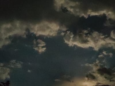 The image shows a cloudy sky at dusk or dawn, with dark and light gray clouds dominating the frame. There is a faint yellow glow on the horizon, suggesting the presence of the sun. In the lower left corner, the dark silhouette of tree branches is visible. The scene appears to be an outdoor environment, possibly a park or a residential area, in the city of Qargaly, Kazakhstan. There are no people or specific objects clearly identifiable.