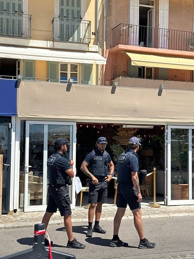 Three men wearing dark blue shirts, vests with "ASVP" written on the back, dark shorts, and baseball caps are standing on a cobblestone sidewalk in front of a storefront. The storefront has large glass windows and doors, and inside, there are displays of what appears to be home decor. The men appear to be security personnel. One man on the left is looking to his left, another in the center is looking forward, and the man on the right is looking to his right. The building behind them has multiple floors with balconies and shutters. The overall setting suggests a sunny day in a Mediterranean town. The presence of "ASVP" on their vests indicates they are municipal police officers in France, and the style of the building and storefront is consistent with the architecture found in coastal towns like Saint-Tropez.
