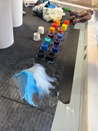 The image displays a tattoo artist's workstation located in Opglabbeek, Belgium. A dark, textured mat covers a section of the work surface, which is bordered on the right by a light-colored table covered with clear plastic sheeting. Seventeen small, clear plastic ink caps are arranged linearly on the dark mat, containing tattoo inks in various colors including black, dark blue, light blue, teal, purple, red, orange, yellow, and white/gold. To the upper left, two tall white cylindrical containers are partially visible. In the upper right background, a green-bodied rotary tattoo machine rests on the plastic sheeting, alongside crumpled paper towels or wipes stained with multi-colored ink residues. On the lower-left area of the dark mat, a substantial quantity of light blue and white creamy ointment or lubricant is spread. The organized setup of inks, tools, and protective coverings indicates an active or recently concluded tattooing session.