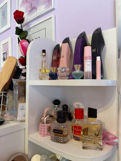 The image shows a close-up view of a white shelf containing various perfume bottles and cosmetic items. On the left side of the shelf, there are several perfume bottles in different shapes and colors, including black, purple, pink, and clear glass bottles. Some of the bottles have labels with the words "Pheramlya Original Perfume Oil" and "IRRESISTIBLE". On the right side of the shelf, there are other perfume bottles, including a gold-colored bottle with the "YSL" logo, a bottle with "Gisada" and "AMBASSADORA" printed on it, a bottle labeled "VALENTINO", and a pink bottle with a handle. Below the shelf, there is a wire basket holding a brush, a comb, and packaged cosmetic items. A red rose and pink tulip appear in the foreground, seemingly placed for decoration. The bottom shelf has more packaged cosmetics, and a makeup compact. Behind the shelf, there is a mirror reflecting the scene, providing a glimpse of a room with framed artwork on a light purple wall. The environment appears to be a bathroom or vanity area. There is no indication of it being in the city of Hassloch, Germany.