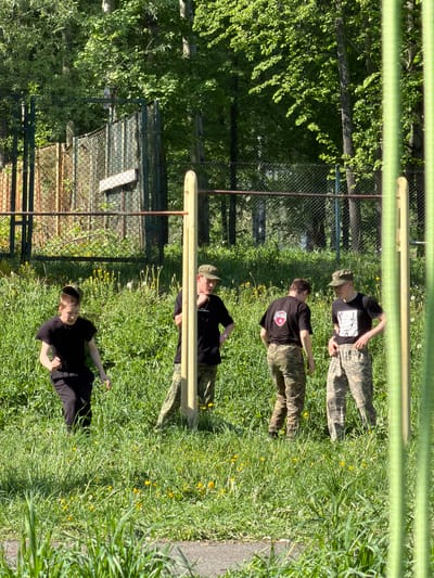 The image depicts a scene in a grassy area, likely a park or training ground, with a wooden obstacle course. Four individuals are present. Three are wearing camouflage pants and black shirts, while the fourth wears all black. One is wearing a camouflage hat. The individuals appear to be either taking a break or awaiting instructions.
In the background, a chain-link fence borders a wooded area. The location is consistent with the city of Oreshniki, Russia, based on the vegetation and infrastructure visible in the image, although the image does not provide a direct location reference. The time of day appears to be midday, based on the lighting and shadows.