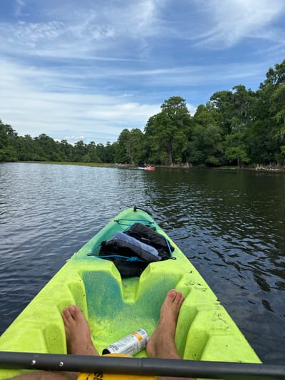 The image captures a first-person perspective from a bright neon green and yellow kayak on a calm body of water, likely a river or lake. The kayaker's bare feet rest on the kayak's textured surface, with a black paddle shaft partially visible on the right. A silver can, possibly bug spray or a beverage, is positioned near the feet, displaying the partial text "CELSIUS" and other small, illegible words. A black dry bag or backpack is secured to the front of the kayak.
The scene unfolds under a broad, light blue sky adorned with wispy white clouds, indicating fair weather. Along the left side of the water, a dense and lush green treeline stretches into the distance, with some trees showing Spanish moss, common in humid environments like Florida. Further down the waterway, faint outlines of other kayaks or small boats can be observed. The water itself is dark and reflects the sky and trees, with gentle ripples disturbing its surface.
The activity depicted is kayaking, suggesting a peaceful outdoor excursion. The time of day appears to be daytime, with clear natural lighting. The overall impression is one of relaxation and connection with nature, consistent with exploring the scenic waterways near Tampa, United States.