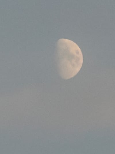 The image shows a moon in the sky, partially obscured by clouds. The moon is a pale yellow color and appears to be in its waning gibbous phase. The sky is a hazy blue, and the clouds are white and wispy. The image is taken from a distance, and there is no way to tell the exact location from the image itself, other than it is outside.