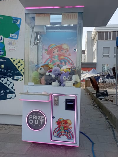 The image shows a claw machine arcade game on a city street. The machine is white and features cartoon characters on the front. It has a clear glass display filled with plush toys. Several stickers are affixed to the side of the machine, including Visa logos and what appear to be credit card numbers. A pink neon light outlines the bottom and a circular "PRIZE OUT" emblem on the machine. The machine is located on a paved walkway next to a street. In the background, there are buildings, some of which appear to be under construction or renovation, with scaffolding and debris visible. A blue hose lies on the ground near the machine. The scene is consistent with an outdoor urban environment in Termez, Uzbekistan, as indicated by the presence of local payment systems and building styles.