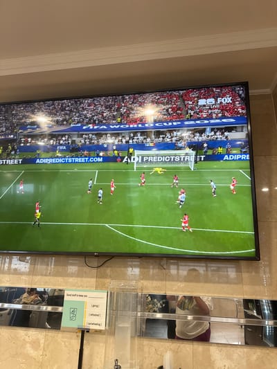 The image shows a large television screen displaying a soccer match, likely the FIFA World Cup 2026, as indicated by the "FIFA WORLD CUP 2026" text on the screen. The broadcast is on FOX. The screen is mounted on a marble wall in what appears to be a public space, possibly a lounge or waiting area. Below the television, there is a mirrored surface and a dispenser with cups. A sign is attached to a stand in front of the dispenser with the text "Good stuff to keep you _______ [going] [refreshed] [cartwheeling]". The flooring appears to be tiled. The image was taken in North Bergen, United States of America.