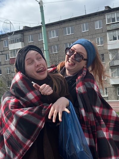 Two young women are huddled together, appearing to be enjoying themselves outdoors. One woman has red hair and is wearing a blue beanie and sunglasses. She has her mouth open, seemingly laughing or smiling. The other woman has dark hair and is wearing a black beanie; she is sticking her tongue out. Both women are wrapped in a red and black plaid blanket. The woman with red hair is holding a crumpled blue plastic bag in her hand. The background features a gray apartment building with multiple windows and an antenna on the roof, suggesting this is in a residential area. The scene takes place in Kachkanar, Russia.