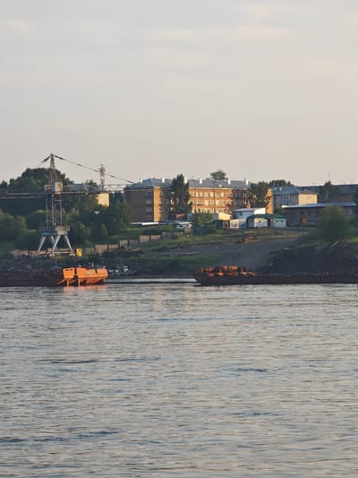 The image depicts a scene along the Angara River in Lesosibirsk, Russia. In the foreground, the calm river water reflects the pale, overcast sky. Two orange barges, weathered and appearing to be under repair or docked for a long period, are prominent on the water's surface. One barge is closer to the viewer, showing its stern with two large, rounded pontoons. The other barge is further away, appearing as a longer, flatter vessel.
The background features a raised riverbank with a mix of industrial and residential elements. A large, industrial crane with a distinctive X-shaped base stands tall on the left side, indicating a port or shipbuilding facility. Behind the crane and along the embankment, several multi-story brick apartment buildings are visible, characteristic of Soviet-era architecture. Smaller structures, including what appear to be sheds or outbuildings, are scattered amongst the buildings. Dense green trees and vegetation line the riverbank and surround the structures. A chain-link fence runs along parts of the embankment. The overall impression is one of a working river town, with signs of both industrial activity and everyday life. There are no visible people in the image.
