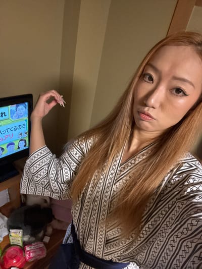 The scene depicts an indoor setting, likely a room within accommodations in Hakuba, Japan. A female individual with long, straight, light brown hair and a light complexion occupies the foreground, looking directly at the camera with a neutral expression and slightly pursed lips. She is wearing a patterned white and black traditional Japanese robe, possibly a yukata, secured with a dark blue obi. Her left arm is bent, hand raised, showcasing her long, pointed, light-colored fingernails. To the left, a small black television screen displays a Japanese program featuring multiple individuals and text. Below the television, on a light wooden surface, rests a small, dark-colored pet, possibly a cat or rabbit, next to various packaged items, including a red container and several packets with Japanese text. The room features plain light brown walls, with vertical darker panels or shadows visible.
