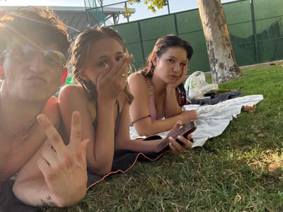 The image depicts three individuals, two females and one male, lying on a patch of grass. The male, positioned on the left, is wearing sunglasses and making a pouting expression, with his right hand raised in a peace sign gesture. The two females are lying next to him. The one in the middle is covering her mouth with her hand, and the one on the right is resting her head on her hand. Both females appear to be wearing swimsuits. They are on a blanket, and a cell phone is visible in the right female's hand. In the background, there is a tree, a tennis court fence, and some bags. The scene appears to be outdoors, with sunlight filtering through the trees. The location is identified as Villa Del Prado, Spain.