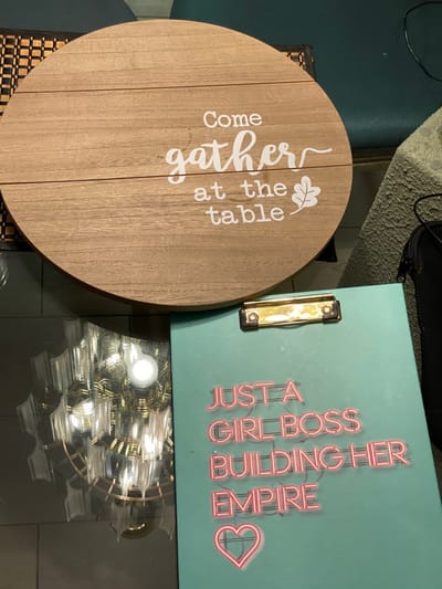 The image shows a round, wooden sign with the words "Come gather at the table" written in white. To the right of the sign is a light blue clipboard with a gold clip, printed with the text "JUST A GIRL BOSS BUILDING HER EMPIRE" in a neon-sign style font. The surfaces of the objects are reflective, showing a reflection of a chandelier-style light fixture. The objects are placed on a dark, possibly glass surface, suggesting a table. The location within Clearwater, United States of America, cannot be determined from the image.