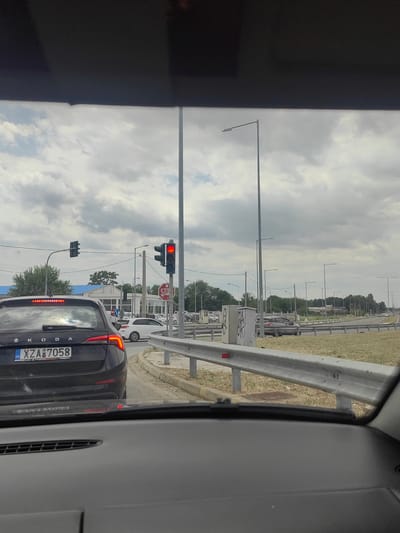 The image shows a view from inside a vehicle, looking forward towards an intersection in Komotini, Greece. The sky is overcast with clouds. In the foreground, the dashboard and the rear of the vehicle in front are visible. This vehicle is a dark-colored Skoda with a Greek license plate reading "XZA 7058".
At the intersection, there are traffic lights. The traffic light on the right is red, indicating vehicles should stop. There is also a red octagonal STOP sign visible. A white car is positioned to the left of the Skoda, and further down the road, a darker car is visible.
Streetlights line the road on both sides. On the left side of the road, there is a building with a blue roof, possibly a gas station. On the right side, there is a metal guardrail separating the road from a grassy area. In the background, there are trees and more buildings. The scene suggests a typical urban environment with traffic control measures in place.