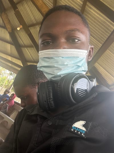 An image captured in Ozuaha, Nigeria, depicts a close-up selfie of an individual positioned in the foreground. This person has dark skin, is looking directly at the camera, and wears a light blue disposable surgical mask covering the nose and mouth. Black over-ear wireless headphones, branded "JBL Wireless," rest on the individual's chest/neck. The person wears a dark-colored, possibly black, collared shirt featuring a subtle patterned texture and a "GLACIER GEAR™" patch with a polar bear logo on the upper right chest.
In the mid-ground, slightly to the left and behind the primary subject, another person with dark skin and short hair is visible, looking downwards. Further in the background, to the left, two additional individuals are seated; one is an older male wearing a light-colored cap and blue attire, and the other is partially obscured, wearing a red hooded garment. The scene takes place under a structure with a corrugated metal roof supported by wooden beams, from which a bare light bulb is suspended. Greenery and a light-colored wall are visible beyond the seated individuals, indicating an outdoor or semi-open setting during daylight hours.
