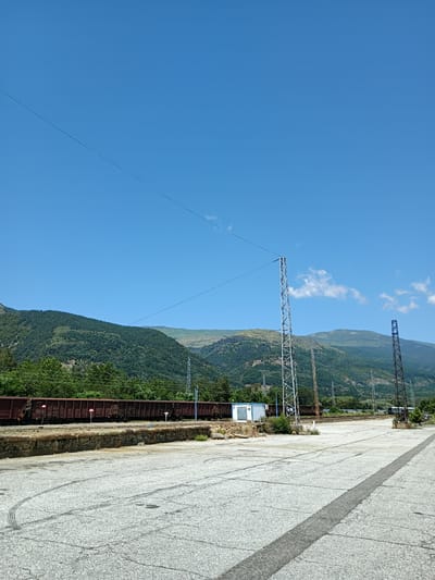 The image depicts an outdoor scene at a train station in Zlatitsa, Bulgaria. The foreground is dominated by a cracked asphalt surface, with tire marks visible. A low stone wall borders a raised area where a long train of freight cars is parked on tracks. The train cars are dark brown and appear to be open-top gondola cars. To the left of the train, a small white modular building stands.

In the midground, several tall metal towers, likely supporting electrical lines or telecommunications equipment, are present. One is a modern lattice structure, while another on the right is older and rusted. Behind the train and towers, a dense forest covers a steep, mountainous terrain. The sky above is clear and bright blue, with a few wispy clouds. The overall impression is that of a working industrial area situated in a natural, mountainous landscape.