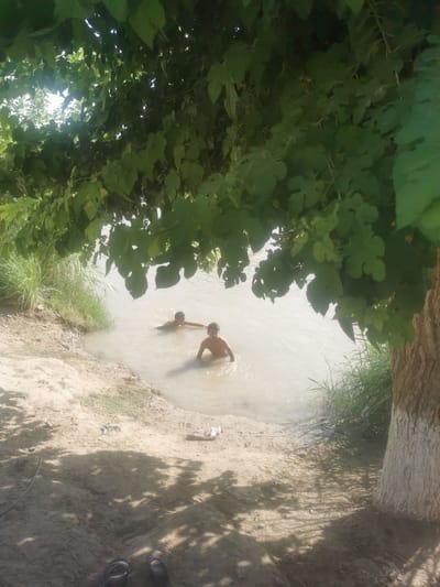 Two young boys are wading in a shallow, muddy river under the shade of a large tree. The boy on the left is partially submerged, with his arms resting on the water's surface. The boy on the right is also partially submerged, with his head and shoulders visible above the water. The riverbank is sandy with some sparse vegetation. In the foreground, a pair of sandals are discarded on the bank. The overall scene suggests a warm, sunny day in a rural area of Jarqurgon, Uzbekistan, where the boys are cooling off in the river.