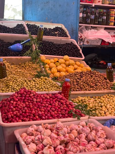 The image captures a colorful array of goods at a market stall, likely in Fnideq, Morocco. Several containers are filled with various types of olives in shades of black, yellow, brown, green, and red. Bunches of fresh parsley are interspersed among the olives. Adjacent to the olives, another container is overflowing with bulbs of garlic. Small plastic bottles containing a liquid, possibly oil or brine, are placed atop some of the containers. A blue plastic scoop rests within one of the olive containers. In the background, a shelf displays rows of bottled goods, adding to the bustling atmosphere of the market.