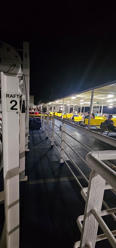 The image depicts the deck of a ferry at night, likely in Split, Croatia, given the context of ferry travel in that region. On the left, a white pillar with a sign reading "RAFT 2" is prominent. Further back on the deck, several passengers are seated on yellow benches under a covered area, illuminated by overhead lights. The deck itself is dark blue with a patterned texture. A metal railing runs along the side of the deck, separating it from the open space. The overall atmosphere is calm, with passengers waiting or resting during the ferry journey.