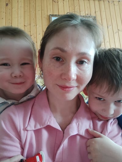 The image depicts a woman with light brown hair and fair skin posing for a selfie with two young children. The woman is centered in the frame, wearing a light pink collared shirt. She has freckles and is smiling slightly. To her left is a young boy with light hair and a wide smile. To her right is another young boy with darker hair looking down. The background is comprised of light brown vertical wooden paneling and a framed picture. A child's hand holding a small red toy car is visible in the bottom left corner. The location is identified as the city of Malaya Purga, Russia.