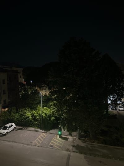 The image shows a night scene in Benevento, Italy, likely in a residential area. In the foreground, there is a paved area with marked parking spots, one of which contains a white SUV. Adjacent to the parking area is a green charging station with a lit green indicator. A tall, thin lamppost stands nearby. Behind the parking area, a dense arrangement of trees and vegetation is visible, illuminated by artificial light. In the background, apartment buildings are partially visible. The sky is dark, with a few faint stars. The overall impression is one of a quiet urban night.