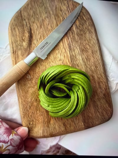 This overhead close-up image captures a culinary arrangement on a wooden cutting board. The main subjects are a knife, a spiral-shaped avocado, and various fabrics.

On the left, a knife with a serrated, silver-colored blade and a light wooden handle rests on the board. The brand "ZARA HOME" is visibly engraved on the blade, followed by smaller, indistinct text.

To the right of the knife, a bright green avocado has been skillfully sliced into thin strips and meticulously arranged into a concentric spiral, commonly known as an "avocado rose." The vibrant green of the avocado stands out against the natural grain of the wooden cutting board. The board itself is light-toned, with visible wood grain and a rounded, organic shape.

The cutting board is placed on a white or light-colored, textured fabric, possibly a tablecloth or napkin. On the far right, a human hand with light-colored fingernail polish is visible, holding a piece of fabric adorned with a floral pattern featuring pink, red, and yellow motifs on a light background.

The scene appears to be indoors, under even ambient lighting, suggesting food preparation or presentation. No specific time of day or location cues for Ros Na Rí, Ireland are discernible from the visual information within the image. The overall impression is one of artful food styling.