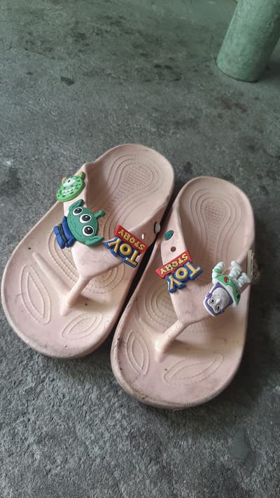 A pair of light pink flip-flops with cartoon characters from Toy Story are displayed on a dark gray concrete surface. The flip-flops are adorned with charms of Buzz Lightyear, a green alien with three eyes, and a "Toy Story" logo. The concrete appears to be outdoors, possibly a patio or sidewalk, with some scattered debris. The scene is static, with no people or animals present. The lighting suggests it is daytime. This image was taken in Kawit, Philippines.