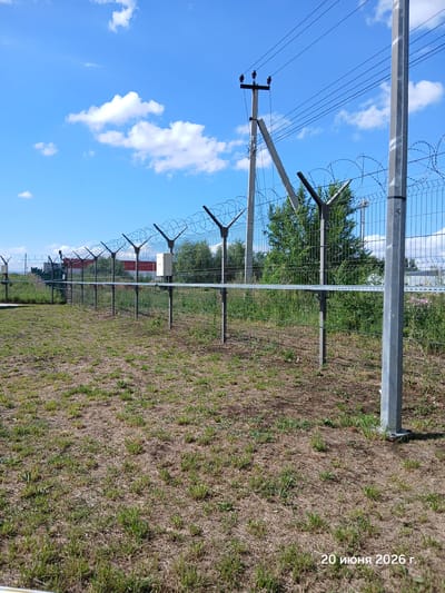 The image depicts an outdoor scene with a clear blue sky and scattered white clouds. In the foreground, a grassy area with dry patches and sparse green grass is visible. A metal fence with barbed wire topping runs horizontally across the image, extending into the distance. The fence is supported by metal posts, one of which is prominently featured on the right side of the frame and has the number "35" painted on it. Behind the fence, there is dense green foliage and trees. In the background, a red and white building can be seen, along with utility poles and power lines. The overall setting appears to be an industrial or perimeter area. A date stamp in the lower right corner reads "20 июня 2026 г." (June 20, 2026).