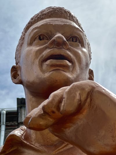 This is a low-angle, close-up photograph of a bronze statue of a man's head and outstretched hand, against a cloudy sky. The statue is a reddish-brown color and appears to be made of bronze. The man's face is upturned, with his mouth slightly open as if speaking or shouting. His right hand is prominently featured, extended towards the viewer. In the background, a building with pipes and a brick facade is visible. The statue is located in the city of Staryi Aidar, Ukraine.