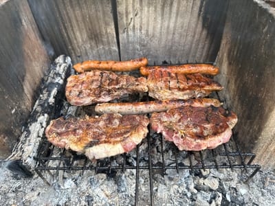 The image shows a close-up of a grill with food cooking on it. There are four pieces of meat (likely pork chops or steaks) and three sausages on the grill. The grill is placed over hot coals or ash, and there is smoke rising from the food and the coals. The grilling is happening in what appears to be a built-in barbecue structure with wooden walls on three sides. The environment suggests an outdoor setting. The location given is Mirkovo, Bulgaria.
