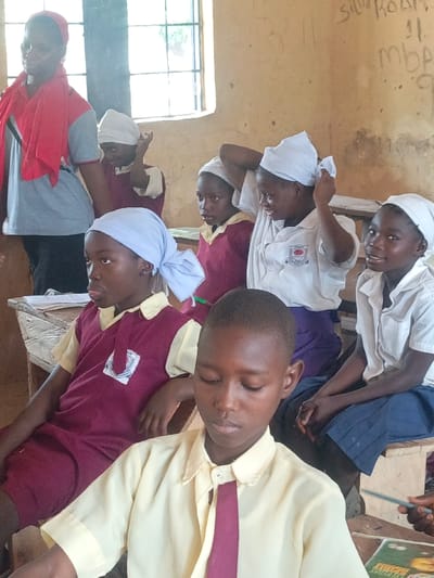 This image depicts an indoor scene within a classroom in Mada, Nigeria. The frame includes one adult female and seven children, appearing to be students. The adult female, standing on the left side, wears a light grey top with black trim and a red garment draped around her shoulders and head. She observes the students. The children are seated on wooden benches and at wooden desks.

Student attire varies: some girls wear maroon tops with cream-colored collars and white headscarves; others wear white button-down shirts, dark blue skirts, and white headscarves. One girl in a white shirt and dark skirt is actively adjusting her white headscarf with both hands. In the foreground, a boy wears a cream-colored collared shirt and a maroon tie, with his gaze directed downwards. To his left, a girl in a maroon uniform and a light blue headscarf is seated at a desk containing papers.

The classroom features light tan walls, with a multi-pane window in the upper left allowing significant natural light. The wall on the right exhibits faded graffiti and writing, including "SILA Room" and "mbp" along with numbers. A pencil and a colorful book are partially visible on the desk in the foreground.
